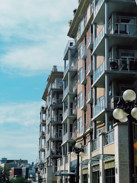 A modern multi-story apartment building with glass balconies under a clear blue sky. The building is made of brick and concrete, with lamp posts visible in the foreground.