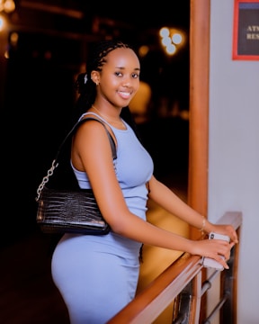 A woman with braided hair wearing a sleeveless blue dress is standing indoors, leaning against a railing while holding a phone. She has a black shoulder bag and is smiling warmly in a softly lit setting.