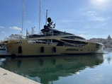 A luxurious yacht is docked at a marina, featuring a sleek and modern design with a shiny metallic gold exterior. In the background, other yachts and boats are visible, along with a shoreline dotted with buildings under a clear blue sky.