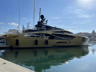 A luxurious yacht is docked at a marina, featuring a sleek and modern design with a shiny metallic gold exterior. In the background, other yachts and boats are visible, along with a shoreline dotted with buildings under a clear blue sky.