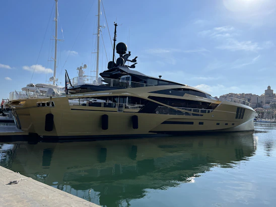A luxurious yacht is docked at a marina, featuring a sleek and modern design with a shiny metallic gold exterior. In the background, other yachts and boats are visible, along with a shoreline dotted with buildings under a clear blue sky.