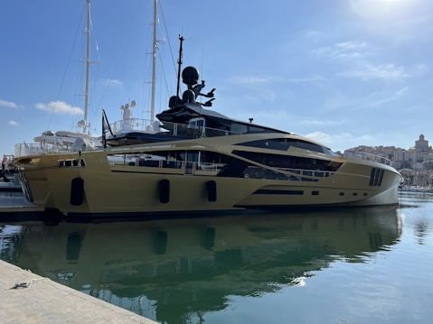 A luxurious yacht is docked at a marina, featuring a sleek and modern design with a shiny metallic gold exterior. In the background, other yachts and boats are visible, along with a shoreline dotted with buildings under a clear blue sky.