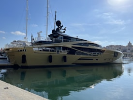 A luxurious yacht is docked at a marina, featuring a sleek and modern design with a shiny metallic gold exterior. In the background, other yachts and boats are visible, along with a shoreline dotted with buildings under a clear blue sky.