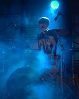 A young person is playing the drums on stage, surrounded by a blue haze or smoke. They are wearing a black T-shirt with a band graphic and are singing into a microphone. The lighting is dramatic, with a prominent spotlight creating a moody atmosphere.