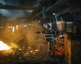 A worker wearing protective gear and a red helmet is managing a rod near a glowing furnace or large fire inside an industrial setting. The environment is filled with smoke and dim lighting, illuminating piles of metal scrap and heavy machinery in the background.