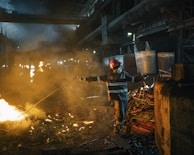 A worker wearing protective gear and a red helmet is managing a rod near a glowing furnace or large fire inside an industrial setting. The environment is filled with smoke and dim lighting, illuminating piles of metal scrap and heavy machinery in the background.