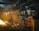 A worker wearing protective gear and a red helmet is managing a rod near a glowing furnace or large fire inside an industrial setting. The environment is filled with smoke and dim lighting, illuminating piles of metal scrap and heavy machinery in the background.