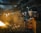 A worker wearing protective gear and a red helmet is managing a rod near a glowing furnace or large fire inside an industrial setting. The environment is filled with smoke and dim lighting, illuminating piles of metal scrap and heavy machinery in the background.
