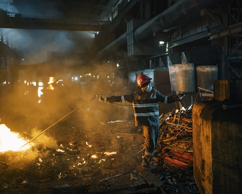A worker wearing protective gear and a red helmet is managing a rod near a glowing furnace or large fire inside an industrial setting. The environment is filled with smoke and dim lighting, illuminating piles of metal scrap and heavy machinery in the background.