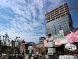 A modern urban area with a tall, partially constructed building in the background featuring glass windows. In the foreground, there are several stacked and creatively decorated shipping containers with vibrant, artistic designs. Some trees and plants are present, adding greenery to the area. A clear blue sky with scattered clouds creates a bright atmosphere.