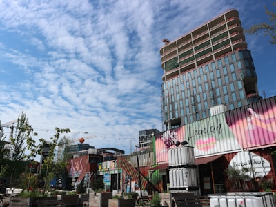 A modern urban area with a tall, partially constructed building in the background featuring glass windows. In the foreground, there are several stacked and creatively decorated shipping containers with vibrant, artistic designs. Some trees and plants are present, adding greenery to the area. A clear blue sky with scattered clouds creates a bright atmosphere.