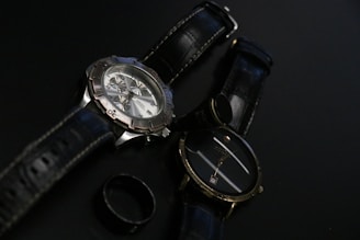 Two wristwatches with black leather straps are placed on a dark surface along with a black ring. One watch features a metallic casing and a detailed face with multiple dials, while the other has a simpler design with a plain face.