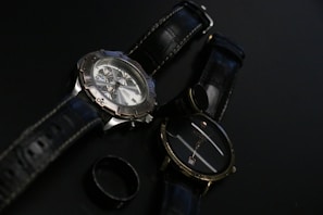 Two wristwatches with black leather straps are placed on a dark surface along with a black ring. One watch features a metallic casing and a detailed face with multiple dials, while the other has a simpler design with a plain face.