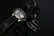 Two wristwatches with black leather straps are placed on a dark surface along with a black ring. One watch features a metallic casing and a detailed face with multiple dials, while the other has a simpler design with a plain face.