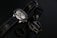 Two wristwatches with black leather straps are placed on a dark surface along with a black ring. One watch features a metallic casing and a detailed face with multiple dials, while the other has a simpler design with a plain face.