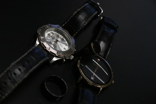 Two wristwatches with black leather straps are placed on a dark surface along with a black ring. One watch features a metallic casing and a detailed face with multiple dials, while the other has a simpler design with a plain face.