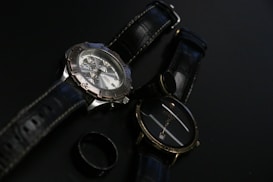 Two wristwatches with black leather straps are placed on a dark surface along with a black ring. One watch features a metallic casing and a detailed face with multiple dials, while the other has a simpler design with a plain face.