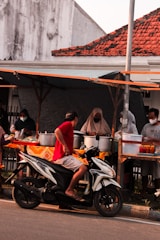 A street food vendor stall with multiple people, including a man on a motorcycle interacting with the vendors. The stall is set up under a makeshift tent, with various dishes, pots, and containers arranged on tables covered in a patterned cloth. The vendors and several customers are wearing masks.
