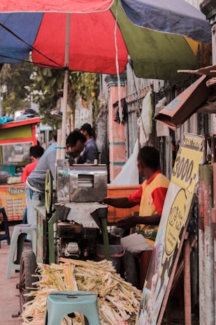 A street vendor sits beneath a colorful umbrella beside a sugarcane juicing machine. There are stalks of sugarcane and signage advertising prices. Various people are visible in the background, creating a bustling street scene.