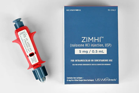 A red and gray injector labeled with the brand ZIMHI lies next to a blue box with printed text indicating it contains naloxone, used for opioid emergencies. The box provides dosage instructions and mentions it is for intramuscular or subcutaneous use.