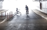 Two cyclists are riding in a dimly lit tunnel, with a bright white light in the background. The person on the left appears to be a child wearing a helmet, while the person on the right is an adult. The road has red and white bollards on the left side, and the tunnel walls are lined with pipes.