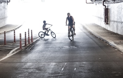 Two cyclists are riding in a dimly lit tunnel, with a bright white light in the background. The person on the left appears to be a child wearing a helmet, while the person on the right is an adult. The road has red and white bollards on the left side, and the tunnel walls are lined with pipes.