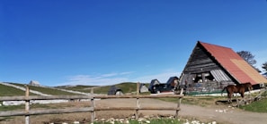 A rural scene featuring several A-frame wooden cabins with distinctively slanted roofs, likely located in a mountainous or hilly area. The structures are scattered across grassy terrain under a clear blue sky. A dirt path runs toward the foreground, bordered by a simple wooden fence. To the right, a horse is standing near one of the cabins.
