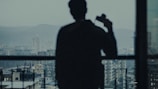A silhouette of a person holding a camera, standing in front of a large window overlooking a cityscape with tall buildings and distant hills under overcast skies.