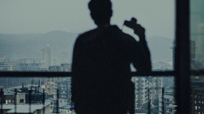 A silhouette of a person holding a camera, standing in front of a large window overlooking a cityscape with tall buildings and distant hills under overcast skies.