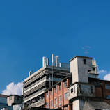 An industrial building complex with a mix of modern and old architecture. The structure features a combination of red brick and concrete surfaces, with visible external piping and protruding antennas. The clear blue sky serves as a backdrop, and a small portion of fluffy white clouds can be seen.