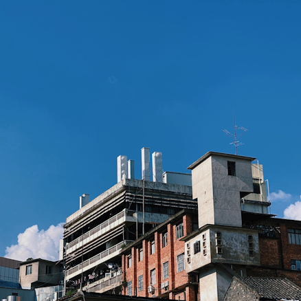 An industrial building complex with a mix of modern and old architecture. The structure features a combination of red brick and concrete surfaces, with visible external piping and protruding antennas. The clear blue sky serves as a backdrop, and a small portion of fluffy white clouds can be seen.