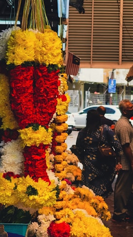 Colorful garlands made of yellow, red, and white flowers are hanging prominently in a market setting. In the background, a few people are present, and a Malaysian flag can be seen, indicating the location. A white car is parked outside on the street.