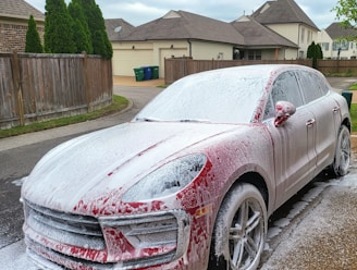 A red car covered in white soap is parked on a driveway near a wooden fence. The background features suburban houses with beige exteriors and brown roofs. Green garbage bins are visible near a garage, and evergreen shrubs add greenery to the scene.