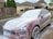 A red car covered in white soap is parked on a driveway near a wooden fence. The background features suburban houses with beige exteriors and brown roofs. Green garbage bins are visible near a garage, and evergreen shrubs add greenery to the scene.