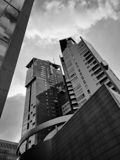 A black and white photograph featuring several modern high-rise buildings seen from a low angle, with a partly cloudy sky in the background. The architecture shows a mix of sleek and angular lines, with reflective glass surfaces and geometric designs.