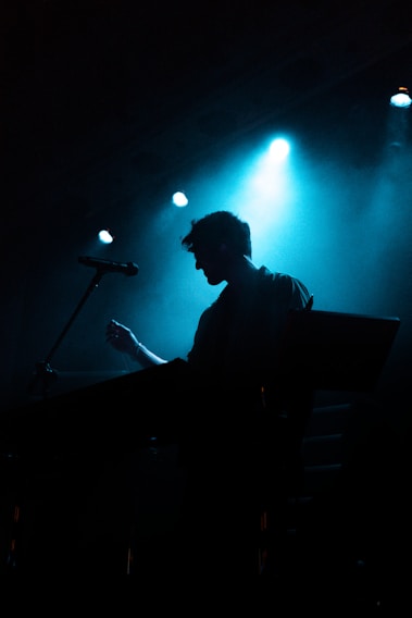 A moody stage shot of Shadowsynth Duo bathed in deep blue and purple lights, the female vocalist immersed in her performance while the male producer adjusts his synthesizers.