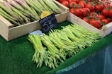 A market stall displays bundles of green asparagus and fresh red tomatoes in wooden crates. Prices are displayed on small signs written in French, with one sign prominently showing the price for asparagus. The produce is arranged neatly on a green artificial grass surface.