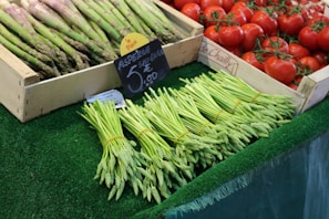 A market stall displays bundles of green asparagus and fresh red tomatoes in wooden crates. Prices are displayed on small signs written in French, with one sign prominently showing the price for asparagus. The produce is arranged neatly on a green artificial grass surface.