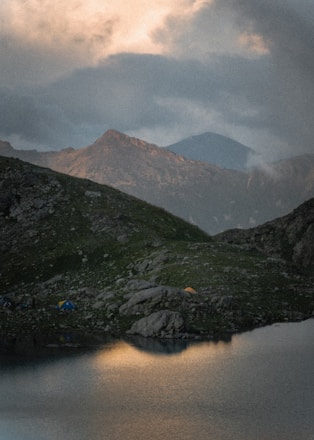 A serene mountain landscape featuring a calm body of water in the foreground, with a grassy hill and rugged terrain. There are tents set up near the water, and mountains rise majestically in the background under a cloudy sky.
