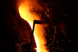 Molten metal pours from a large industrial furnace, illuminating the surrounding area with an intense orange glow. Sparks fly and a silhouetted figure can be seen nearby, engaged in the metalworking process. The scene is dominated by the fiery liquid and dark shadows, creating a stark contrast.
