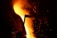 Molten metal pours from a large industrial furnace, illuminating the surrounding area with an intense orange glow. Sparks fly and a silhouetted figure can be seen nearby, engaged in the metalworking process. The scene is dominated by the fiery liquid and dark shadows, creating a stark contrast.