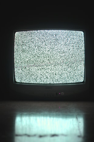 A television displaying static noise on the screen in a dimly lit room. The reflected light from the screen casts a faint glow on the surface in front of it.