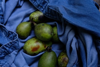 Several green fruits, possibly avocados or pears, are placed on a crumpled piece of blue denim fabric. The fabric creates a textured background that contrasts with the smooth surface of the fruits.