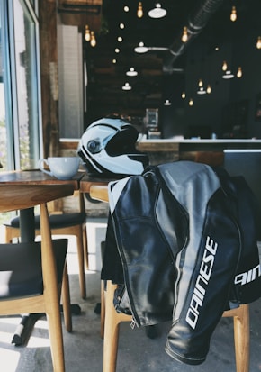 A motorcycle helmet and jacket are placed on a chair in a cozy cafe with wooden furniture and warm lighting. The cafe's interior features exposed ductwork and hanging light fixtures, creating a modern and inviting atmosphere. The jacket is black with white lettering, and the helmet is white with black accents, sitting next to a white coffee cup on the table.
