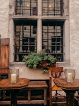 A rustic outdoor setting featuring two wooden tables and matching chairs below a large, multi-pane window. The window has black iron bars and is adorned with a planter filled with lush green plants. Each table has a large glass jar holding a white candle. The wall is textured and painted in a light cream color, enhancing the vintage ambiance.