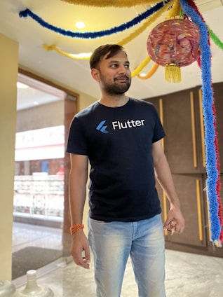 A person wearing a dark t-shirt with the word 'Flutter' stands under colorful hanging decorations. The setting appears to be indoors, with a wooden cabinet and a large window visible in the background.