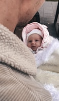 A baby wrapped in warm clothing is being held by an adult. The baby is wearing a pink and white hooded outfit with floral patterns. The adult is dressed in a beige textured jacket. The background includes a paved surface.