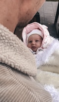 A baby wrapped in warm clothing is being held by an adult. The baby is wearing a pink and white hooded outfit with floral patterns. The adult is dressed in a beige textured jacket. The background includes a paved surface.