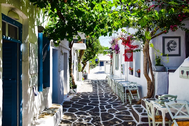 A narrow, charming street in a Mediterranean village with whitewashed buildings and colorful shutters. Plants and vibrant flowers, including bougainvillea, cascade from above, while tables and chairs line the pathway, creating an inviting atmosphere.