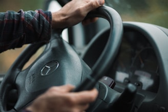 Hands gripping the steering wheel of a vehicle, focusing on the Mercedes-Benz logo prominently displayed at the center. The dashboard and part of the instrument cluster are visible in the background. The lighting appears natural and subdued, hinting at an outdoor setting.